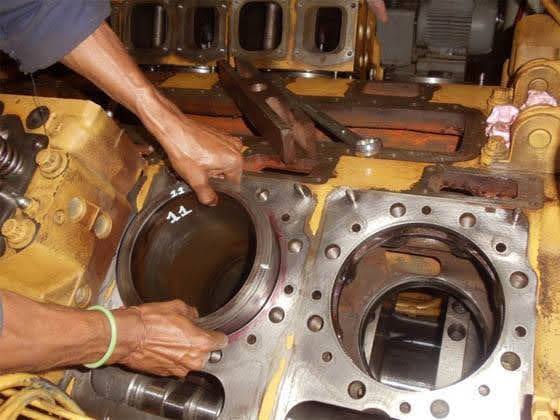 Technician handling a numbered cylinder liner over an open engine block, with bores and head-bolt holes visible during a generator engine overhaul