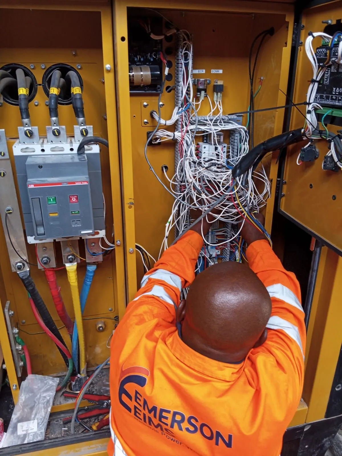 EmersonEIMS technician in a branded hi-vis jacket rewiring the control cabinet of a CAT generator