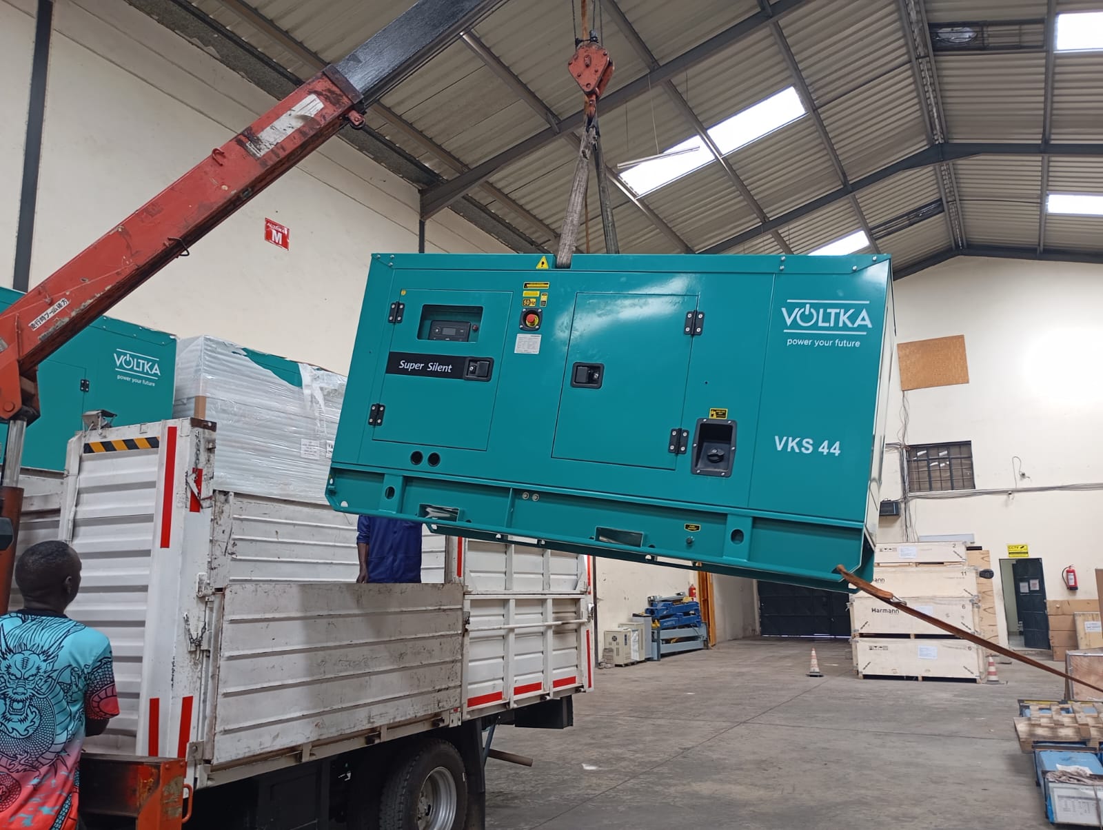 Canopied super-silent generator set lifted by crane inside the EmersonEIMS warehouse, showing the complete acoustic enclosure, access doors and control-panel cut-out