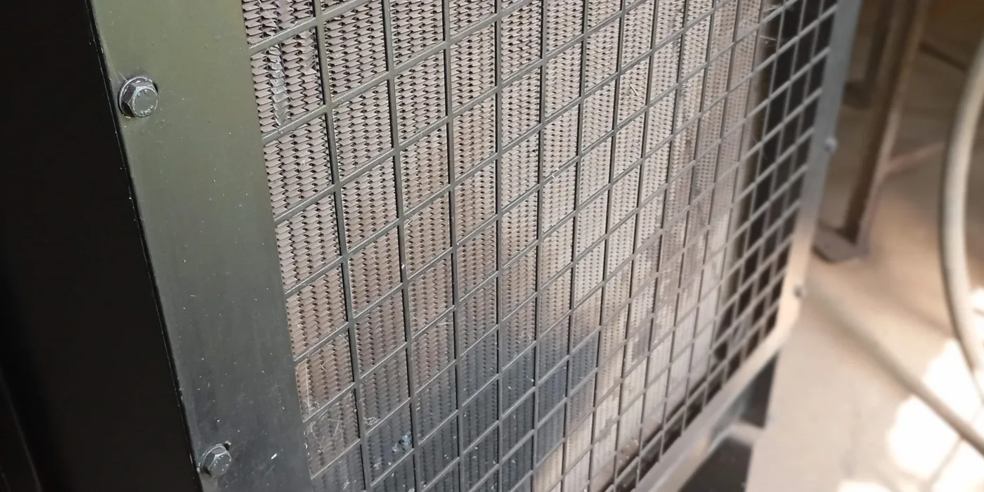 Close-up of a generator radiator fin-and-tube core behind its protective mesh guard, the surface loaded with dust that restricts cooling airflow